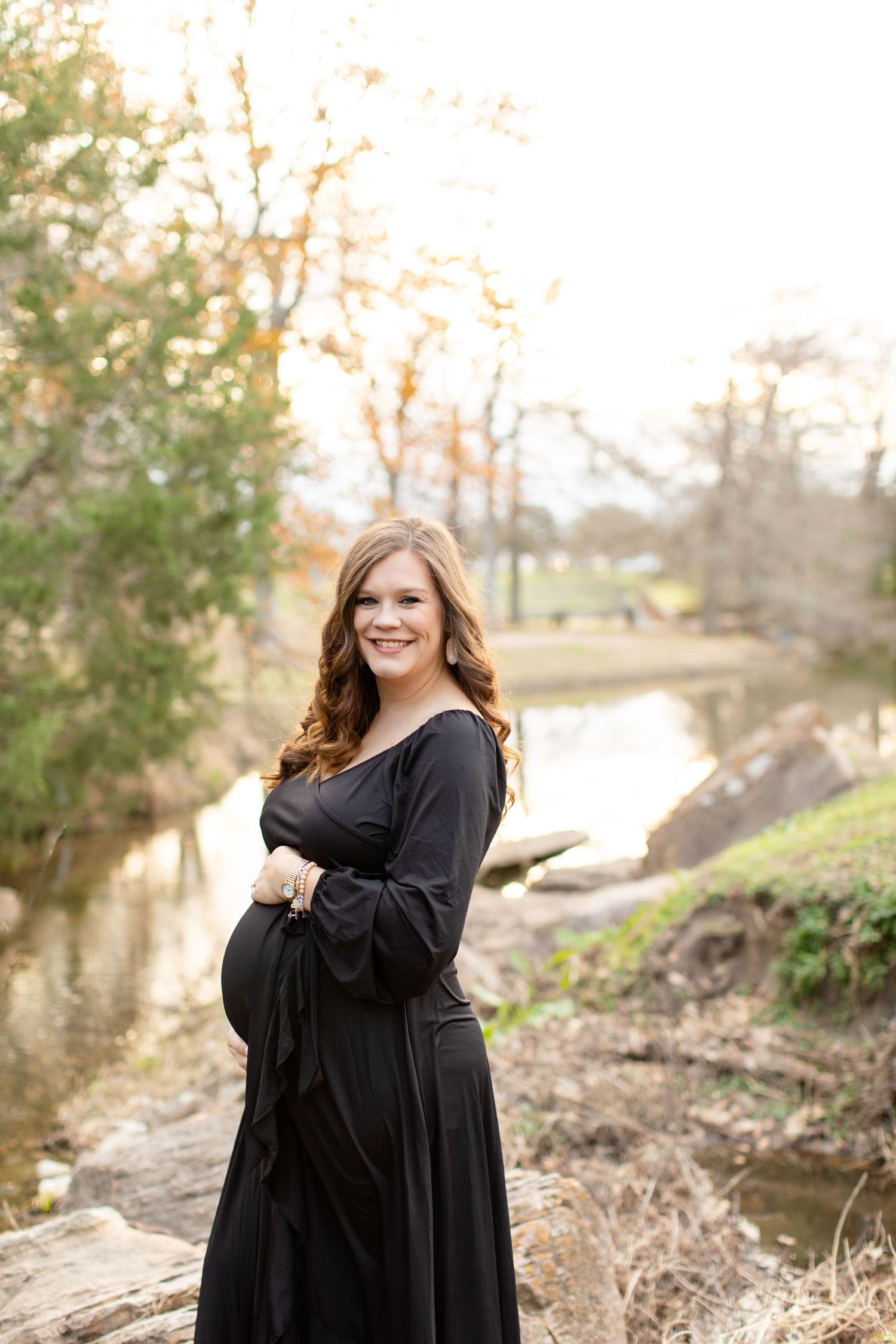 Expectant mother in flowing black dress stands beside creek during golden hour photoshoot.