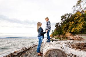 Mom stands on the beach and looks up at her son who stands on driftwood at Lincoln Park, West Seattle.