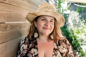 Woman leans against a wooden fence for an outdoor, backyard headshot session wearing a straw hat and floral dress.