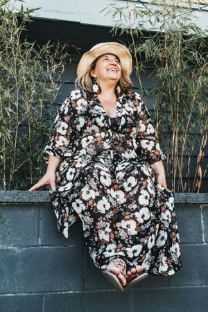 Woman sits on a wall looking up to the sky, wearing a floral dress and a straw hat, next to bamboo plants.