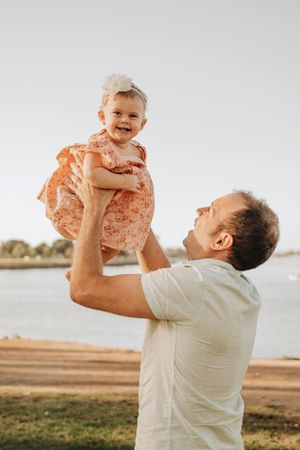 Joyful outdoor photos of adult lifting baby wearing peach dress against waterfront backdrop.