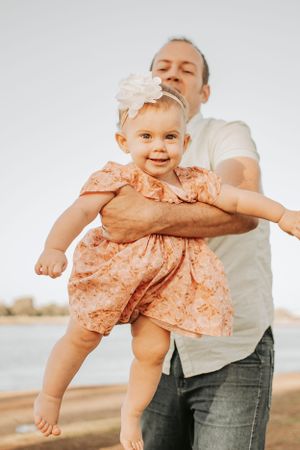 A toddler in a peach floral dress being lifted playfully in the air by an adult against a light background.