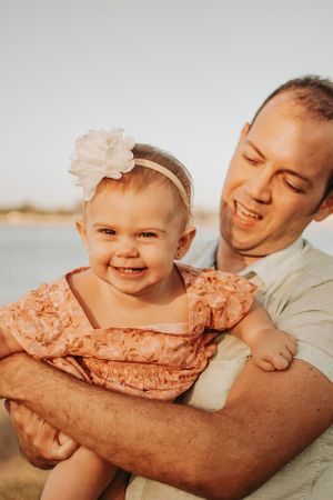 A baby wearing a peach-colored dress smiles brightly while being held at sunset.