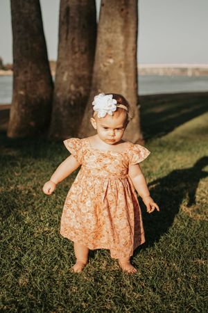A small child in a peachy floral dress stands on grass near palm trees at sunset.