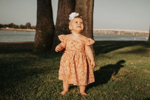 A toddler in a peach dress explores outdoors near tall palm trees during golden hour.
