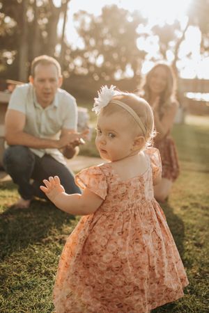 A toddler in a floral dress stands in golden sunlight while adults watch from behind.