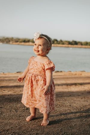 Baby in peach floral dress explores sandy beach during sunset.