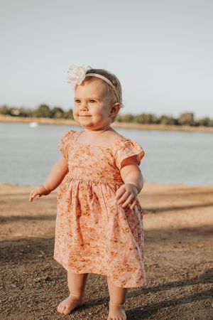 Toddler in peach dress stands on beach during golden hour photoshoot.