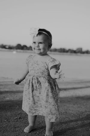 Black and white photo of toddler in dress standing on beach.