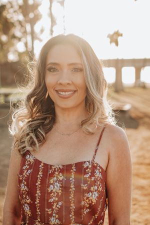 Woman in red floral sundress poses outdoors in warm sunlight during golden hour.