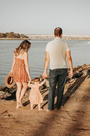 Family walks along rocky shoreline holding hands at sunset near ocean and bridge.