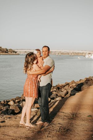 Couple shares intimate moment on rocky beach path with water and bridge in background.