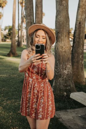 Woman in red floral dress takes selfies and laughs among palm trees.