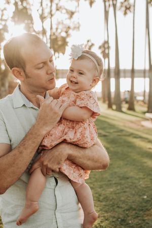 Parent holds and kisses baby wearing pink dress in sunny park setting.