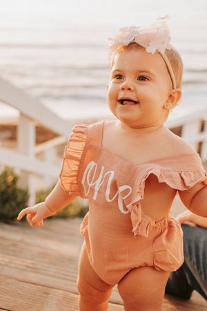 Baby in peachy-pink ruffled romper smiling at beach.