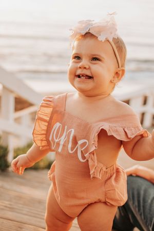 Baby wearing pink outfit with ruffles and 'one' text smiling at beach.