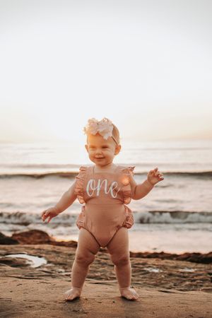 Baby in peach romper standing on beach at sunset.