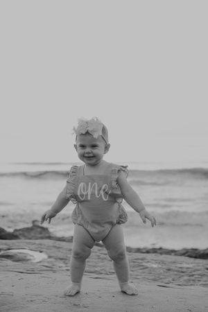A toddler wearing a ruffled 'one' outfit stands on a beach shore in black and white.