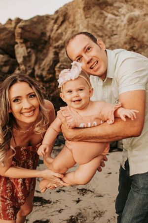 A happy family of three poses together on a beach with rocky cliffs in the background.