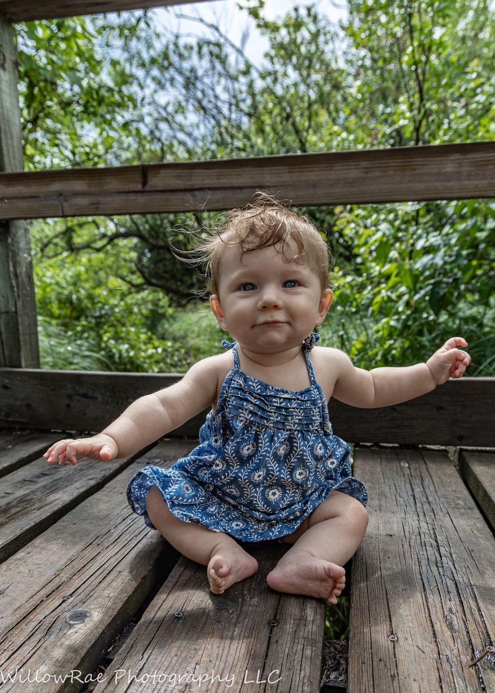 Happy baby in blue patterned dress sitting on wooden deck outdoors.