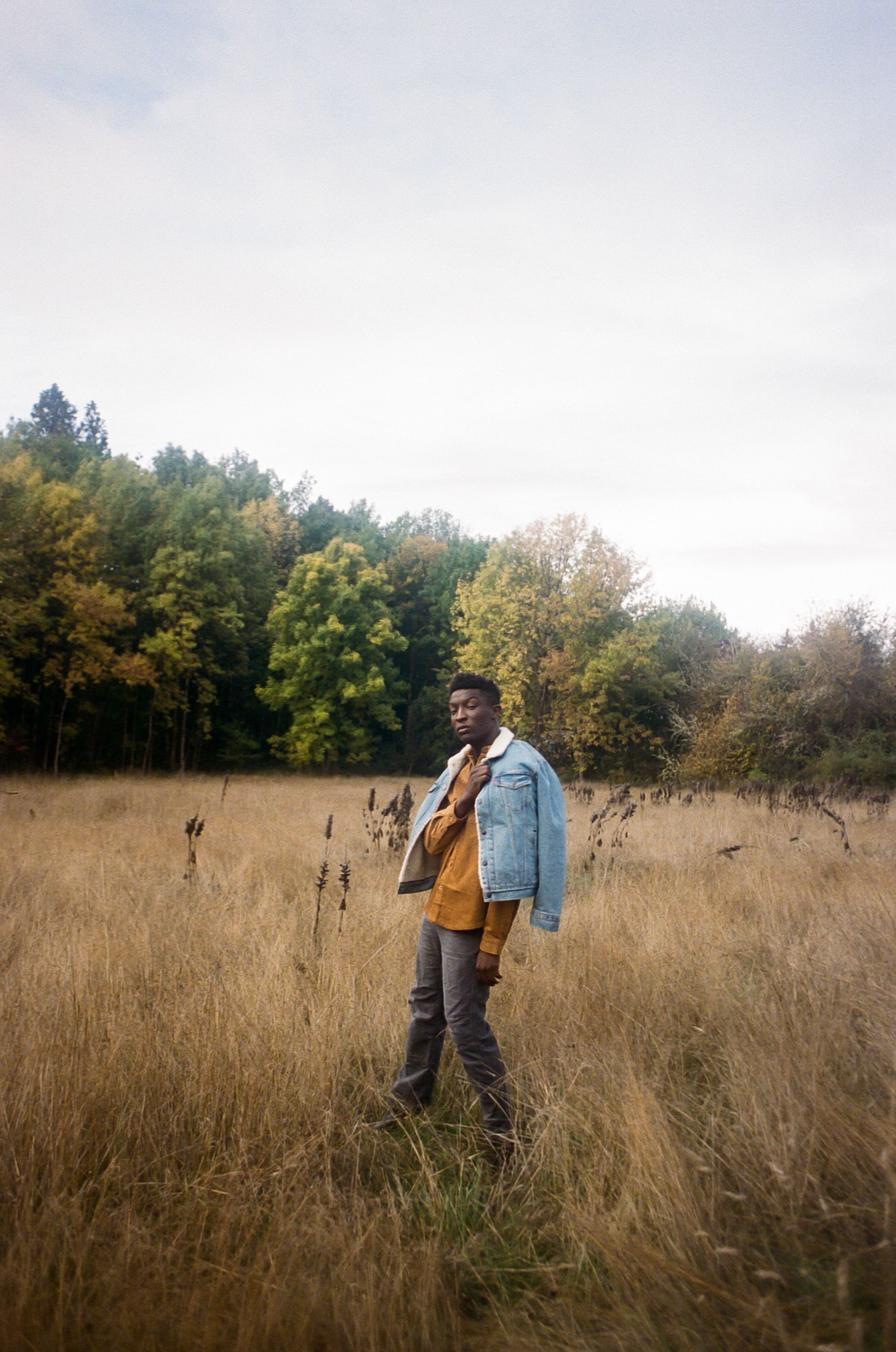 A person stands in a golden autumn field wearing a denim jacket and brown corduroy shirt against a wooded backdrop.