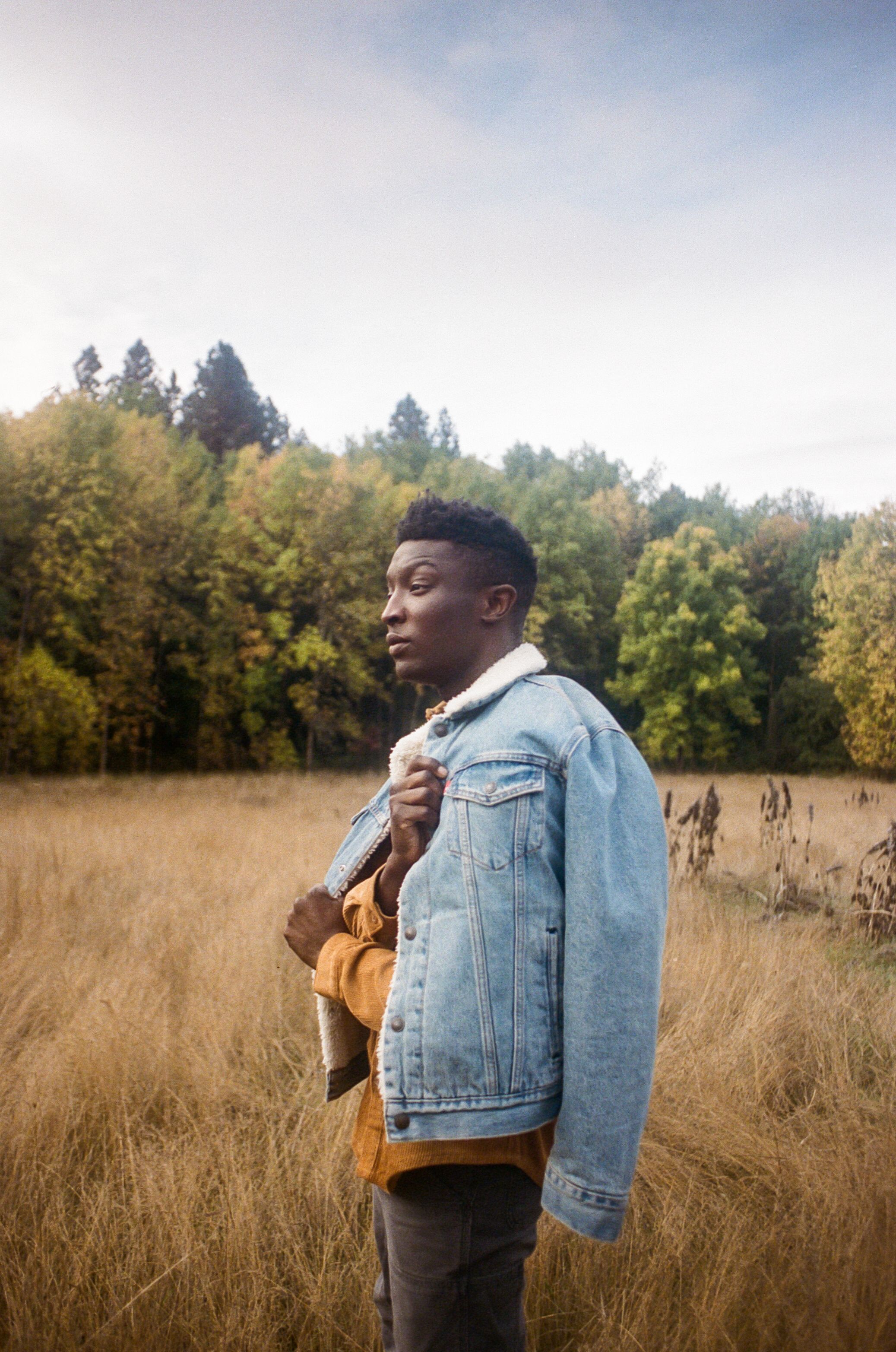 A person in a denim jacket poses thoughtfully in tall grass with autumn trees in the background.