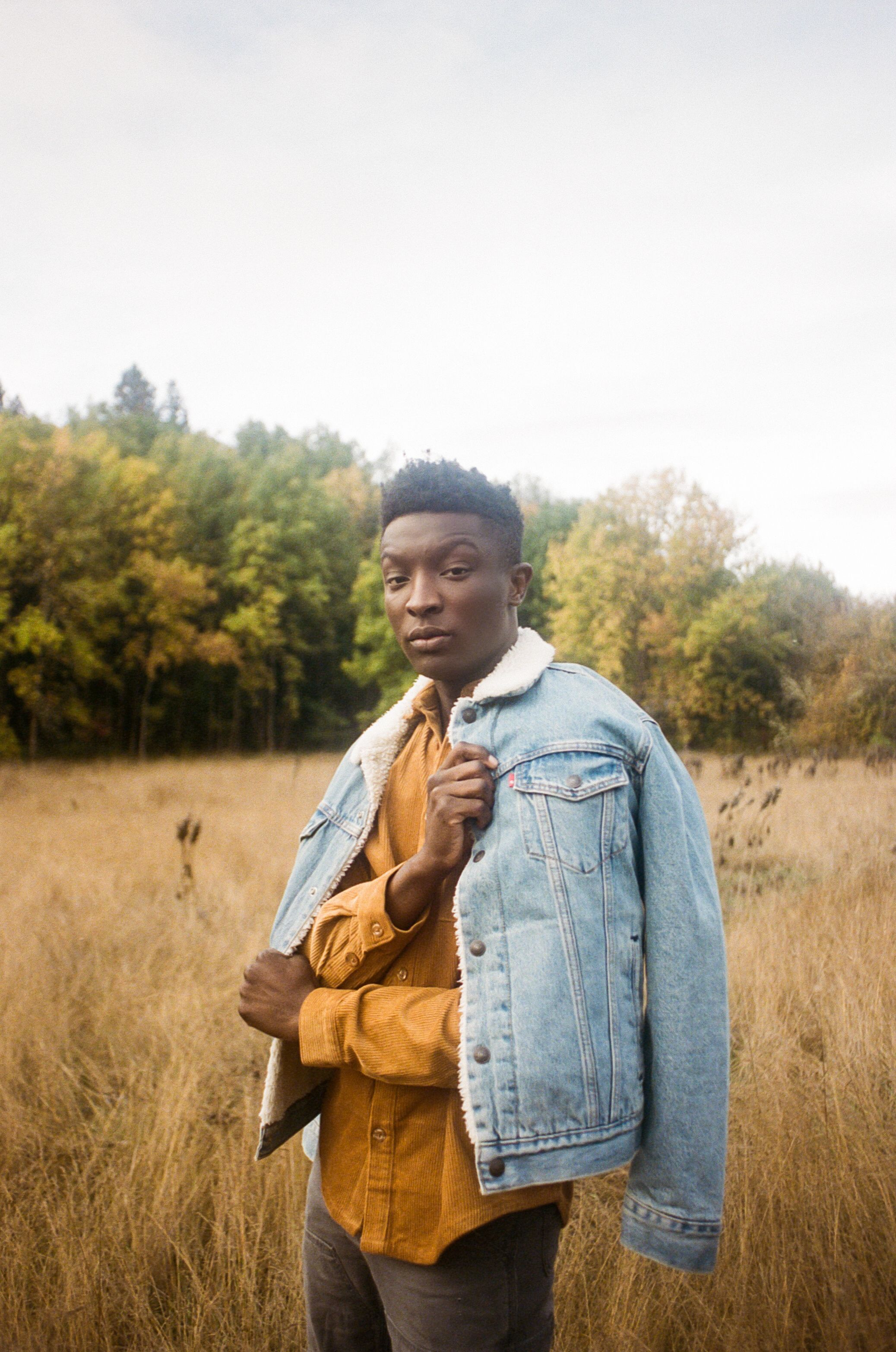 A person wearing layers of denim and brown corduroy stands in a sunlit meadow during fall.