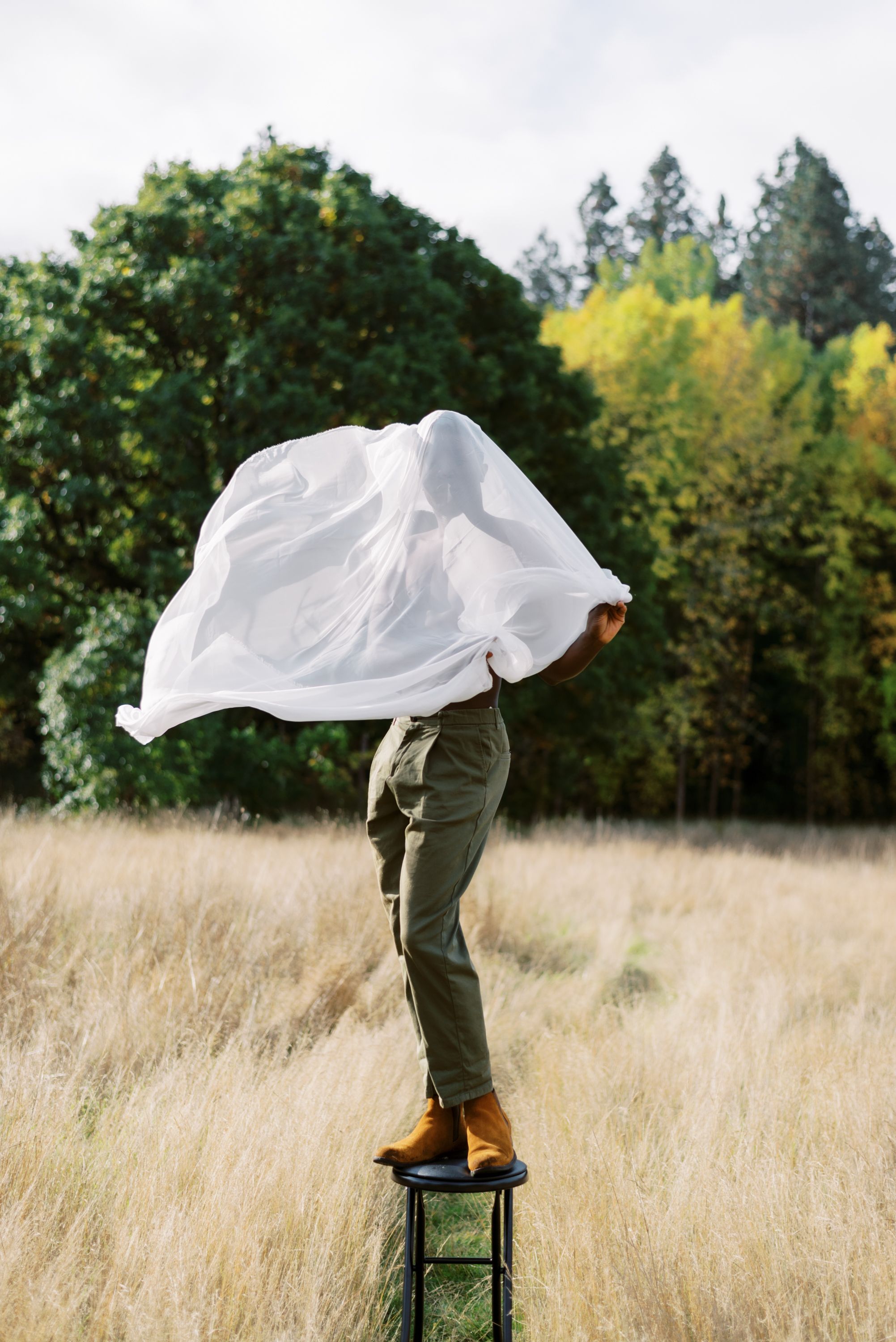 White fabric billows over person balanced on green stool in tall grass field.