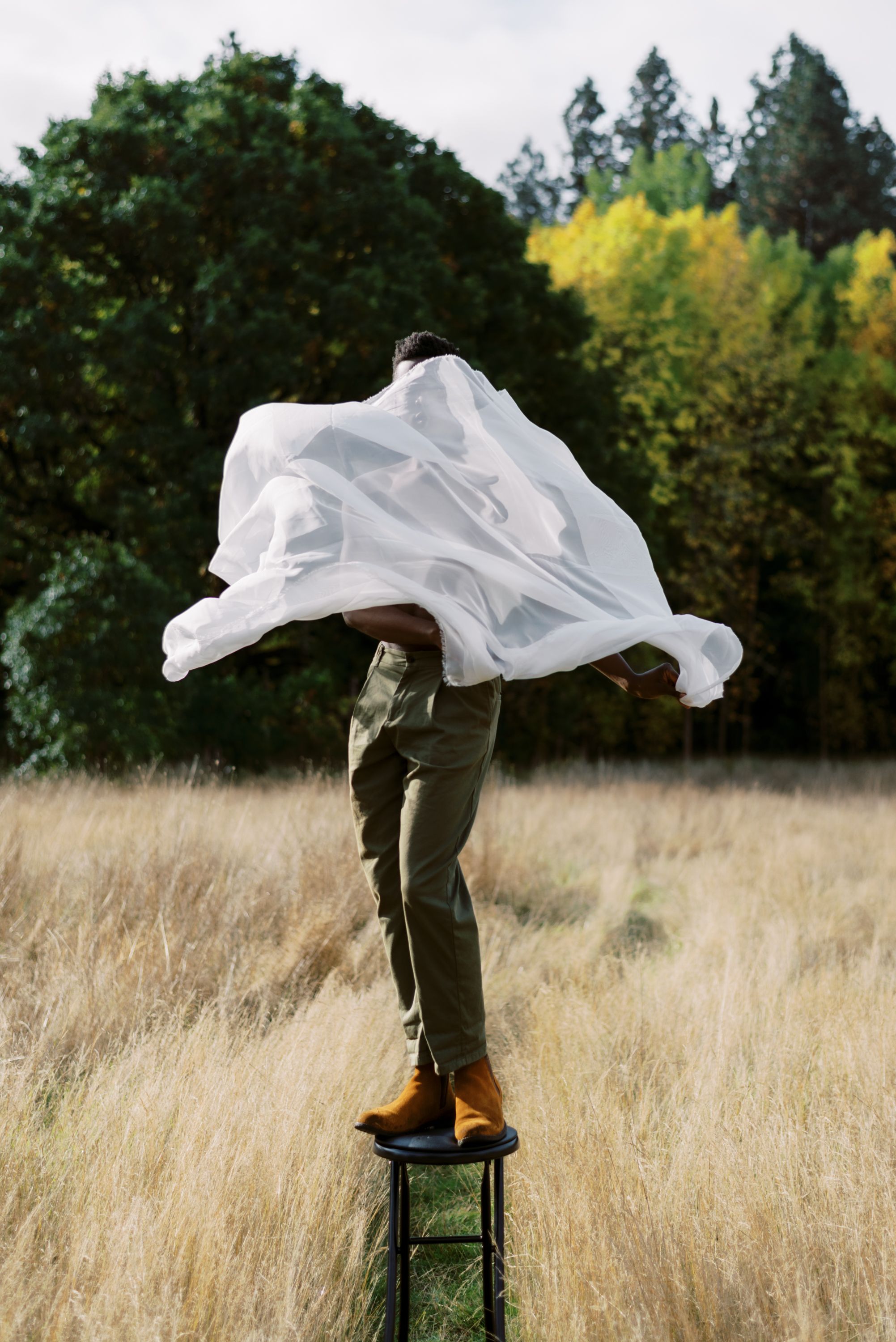 Flowing white fabric catches wind as figure stands on stool in autumn field.