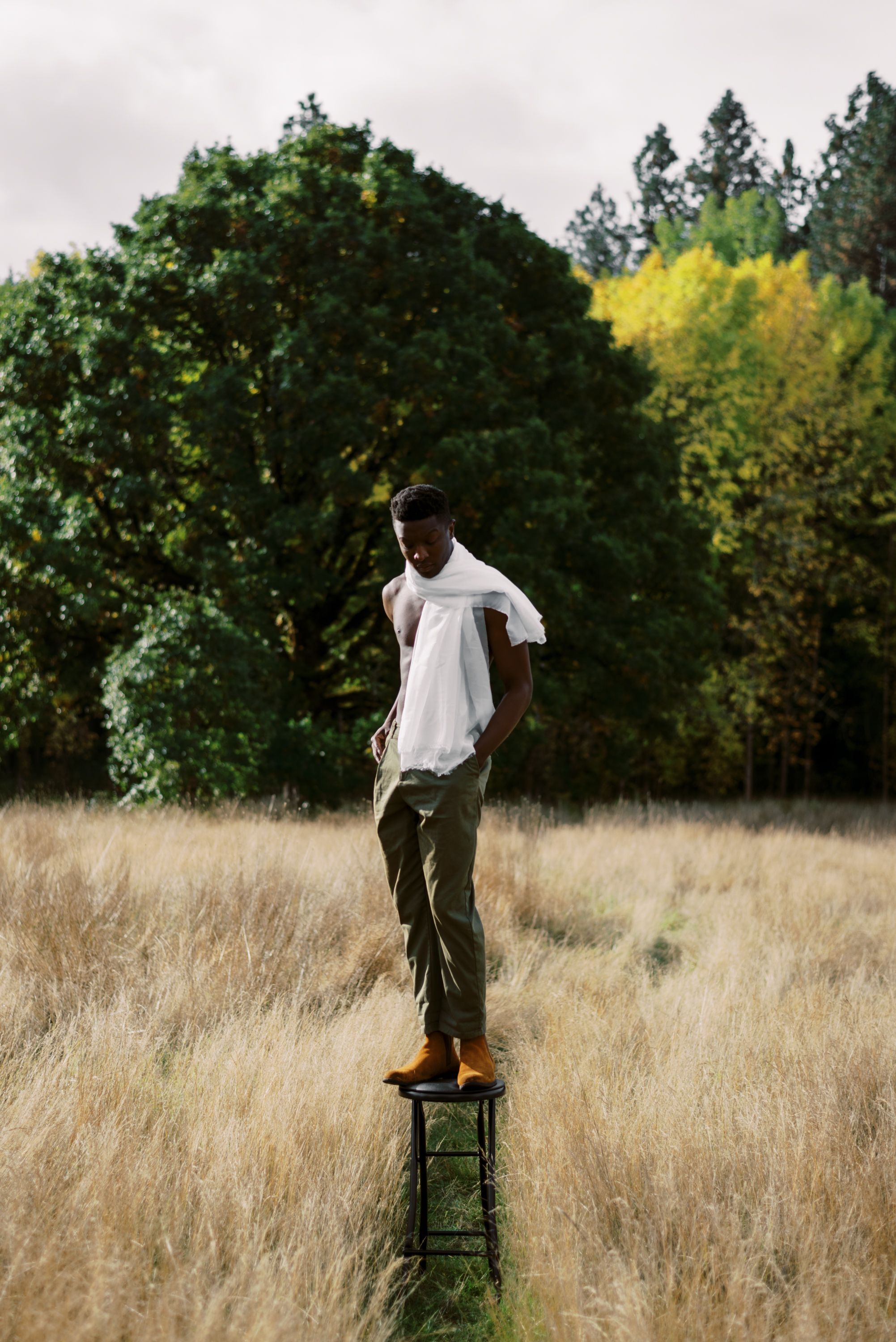 Person in casual attire stands on green stool in tall grass with trees behind.