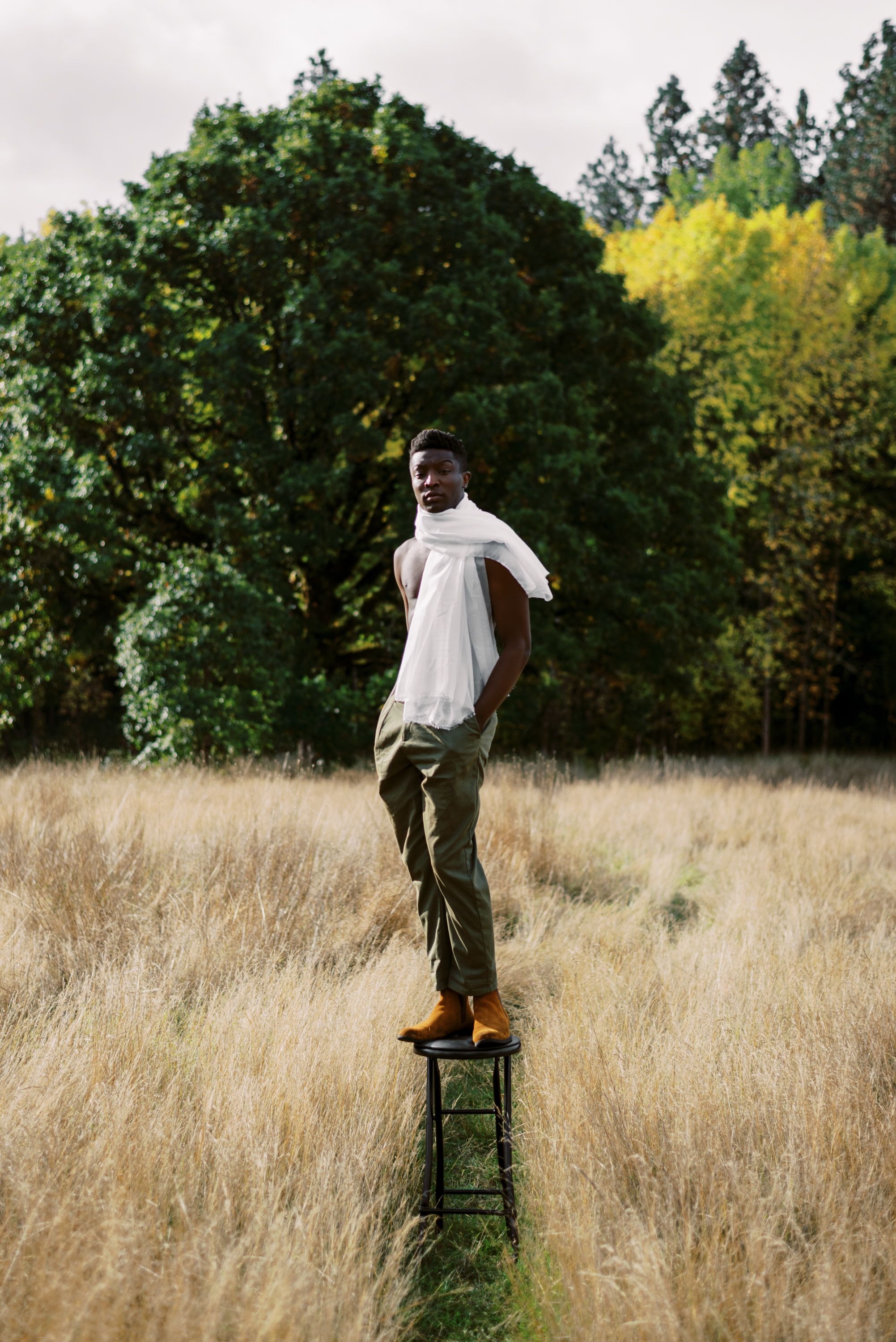 Figure in white shirt poses on vintage stool against autumn forest backdrop.