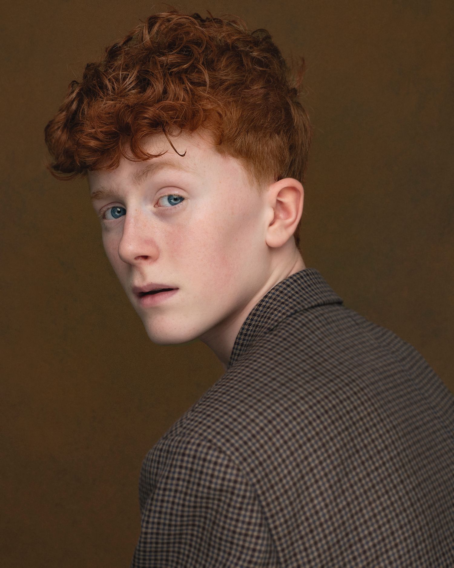 Studio portrait featuring red curly hair and plaid jacket against brown background.