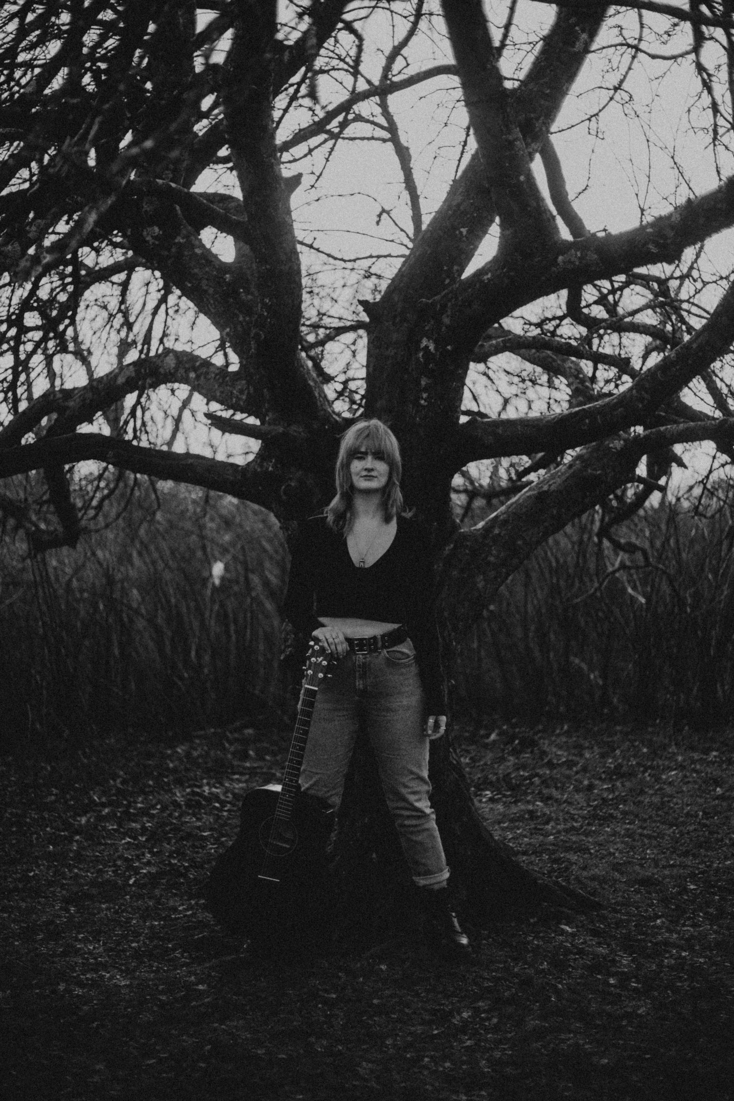 Black and white photo of a joyful figure standing in front of a large bare tree on a moody day.