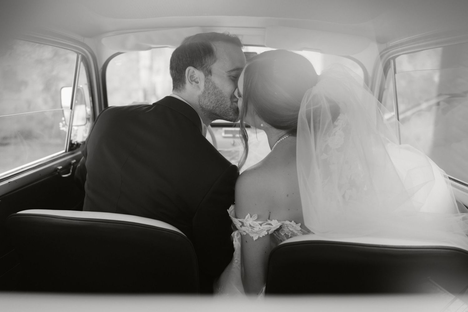 A black and white photo of newlyweds sharing an intimate moment in a vintage car.