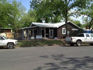 Small brown ranch house with covered porch and parked trucks in front on a tree-lined residential street.