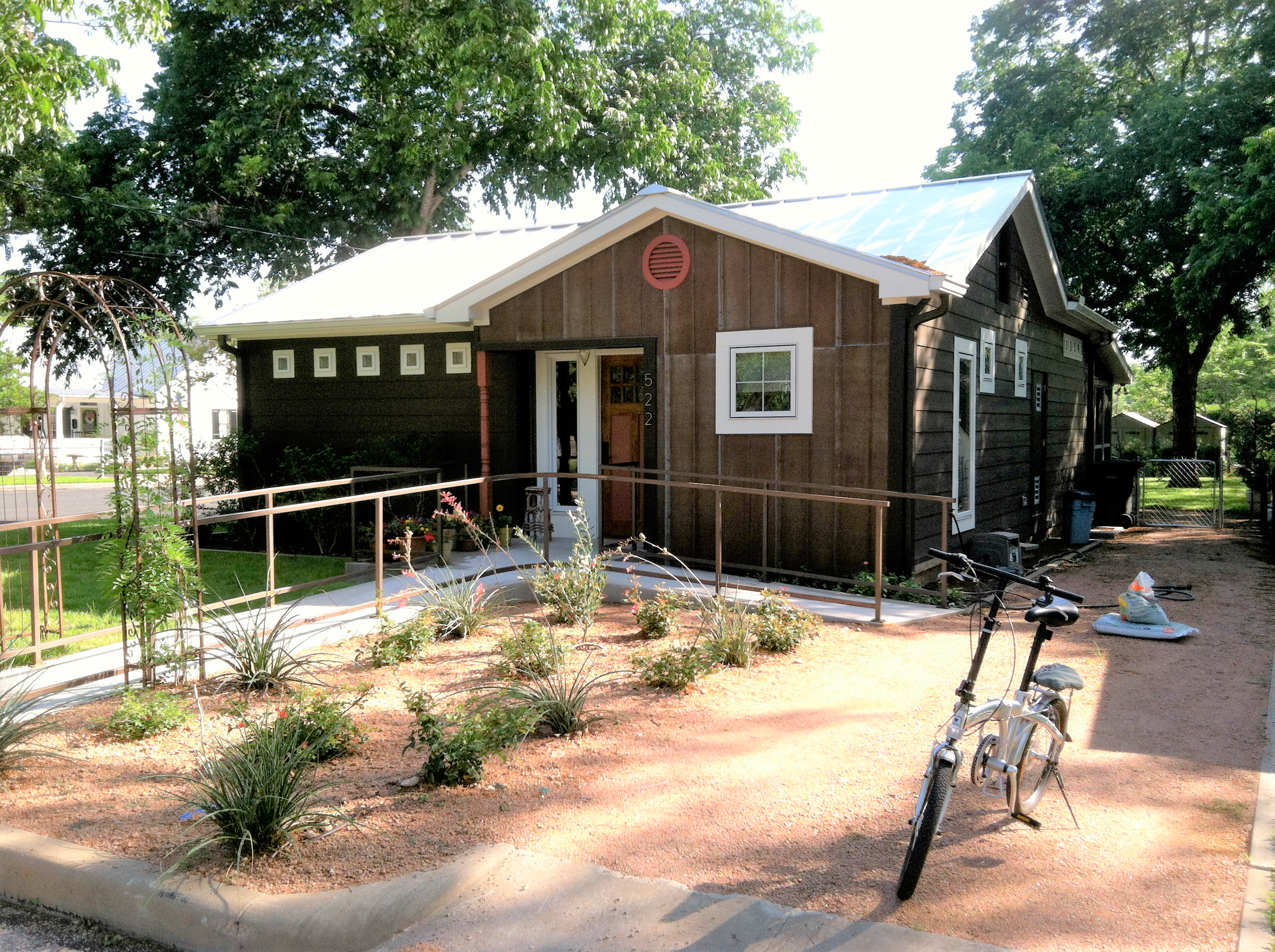 A brown wooden cabin with a wheelchair ramp and desert landscaping sits under shady trees on a sunny day.