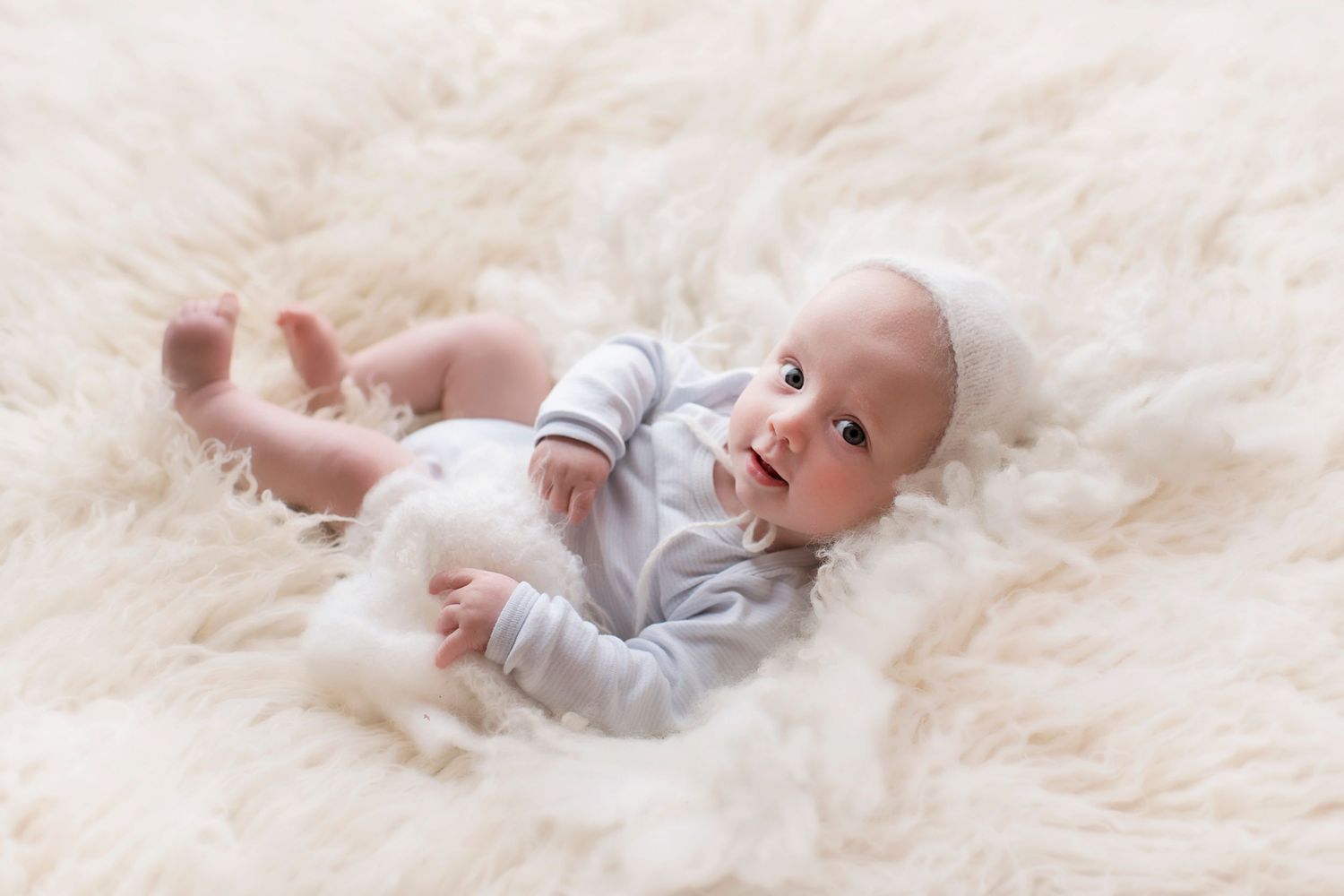 A smiling infant lies on their back on soft white fur wearing light gray clothing.
