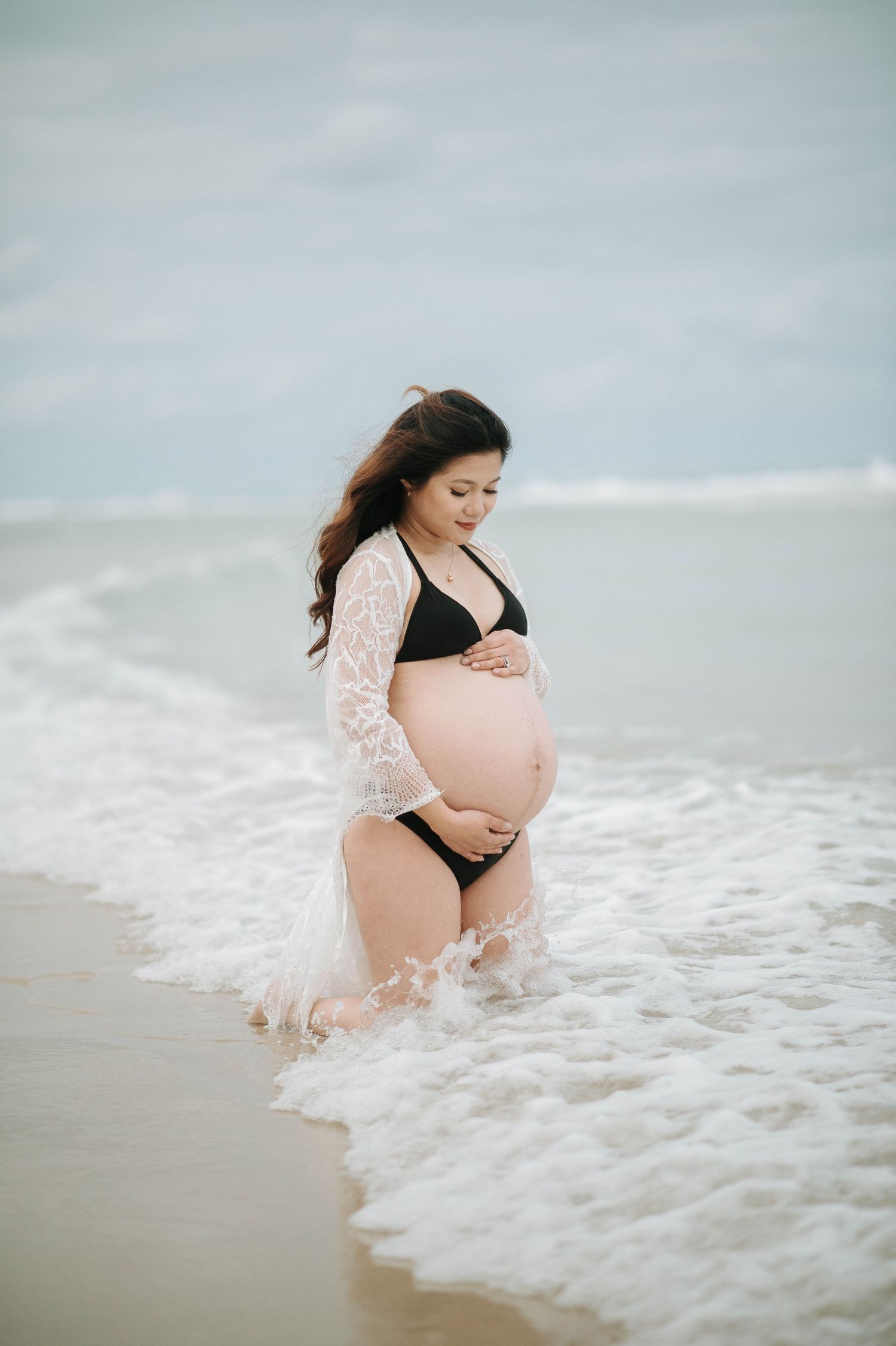 An expectant mother in a lace cover-up and black swimsuit standing in gentle ocean waves.