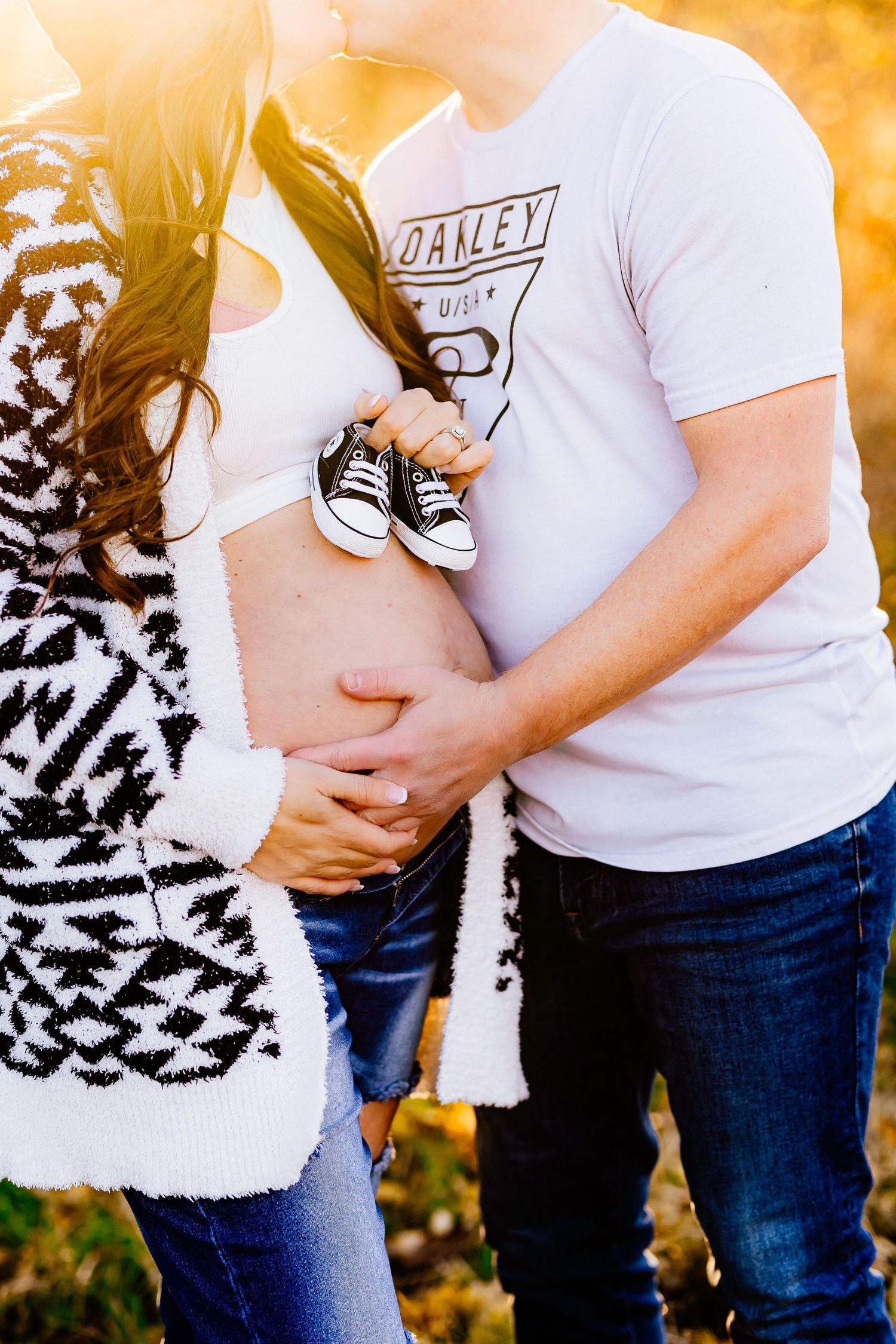 Expectant couple holding tiny baby shoes against sunset backdrop with black and white patterned blanket.