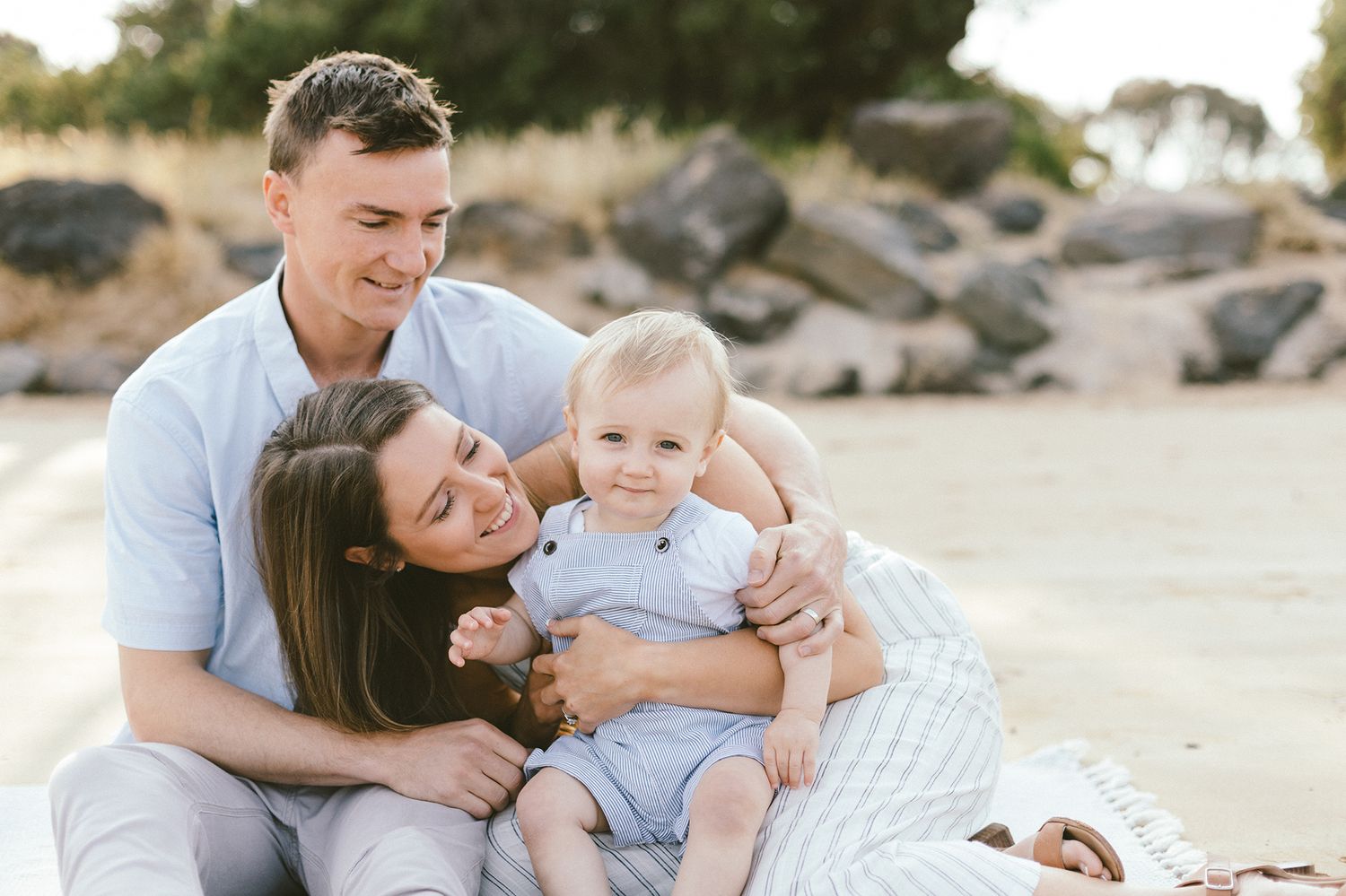 Parents cuddle with baby on beach as sunlight streams through trees.