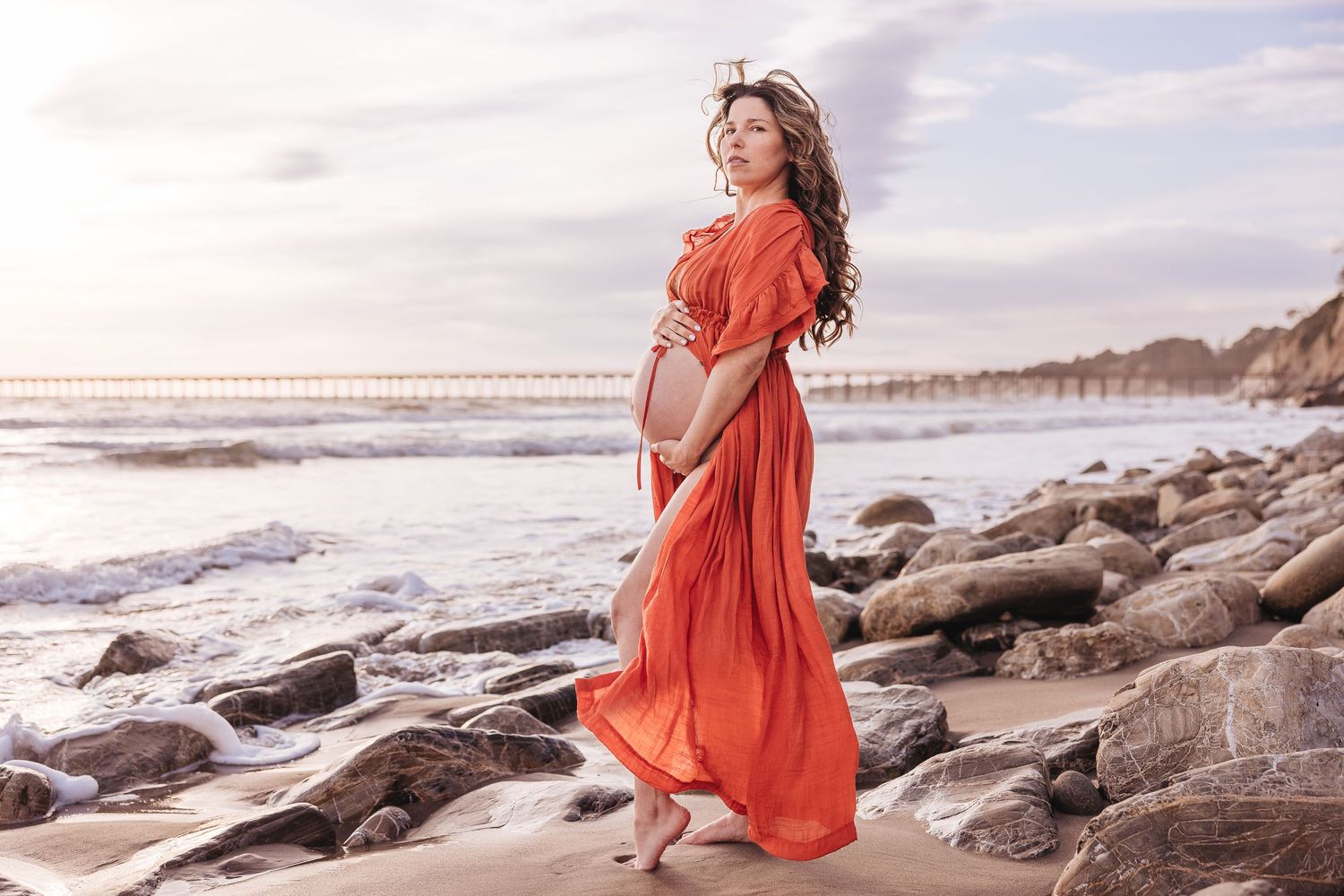 Expecting mother in flowing coral dress poses gracefully on rocky beach at sunset.