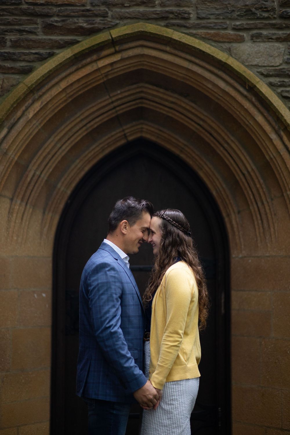A couple shares an intimate moment under a gothic stone archway, with the person in yellow and navy blue suit.