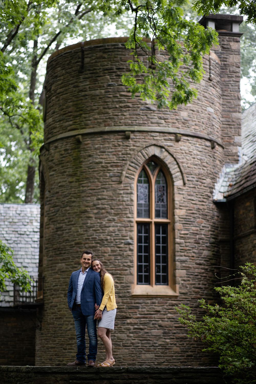 Couple in yellow and blue stand together against a stone castle tower with gothic windows.