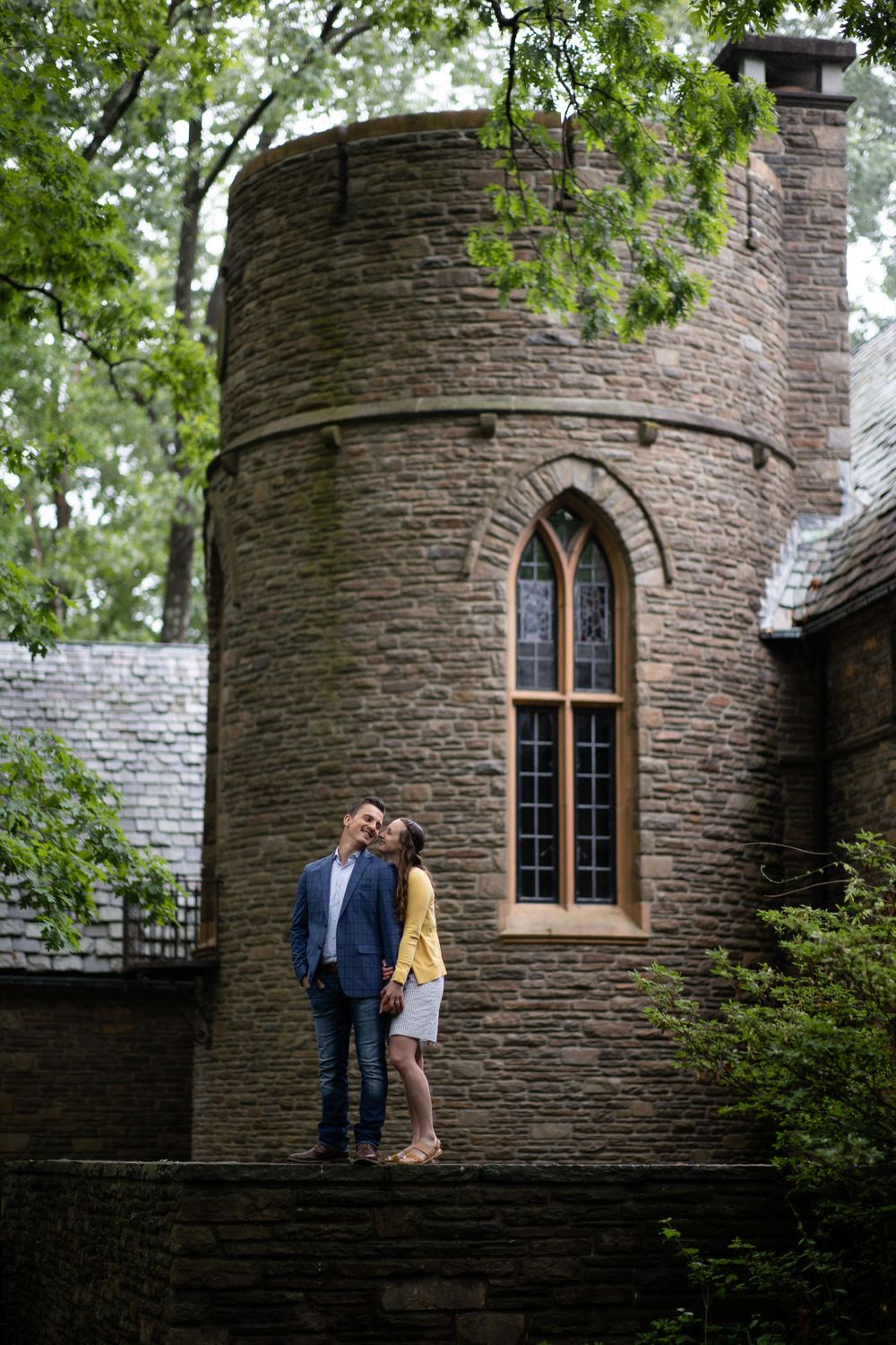 Romantic pair pose on stone steps by a circular tower with arched windows.