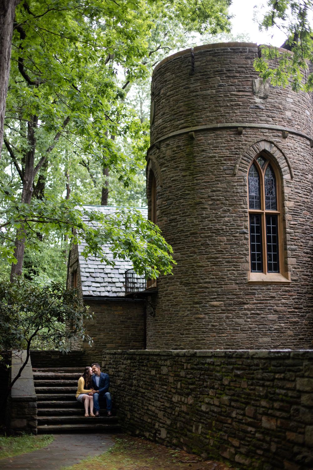 Stone castle tower with gothic window framed by green trees and couple sitting on steps below.