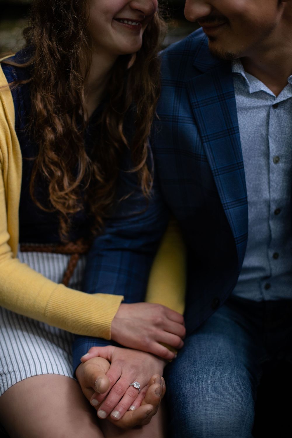 Close-up of clasped hands showing engagement ring, with yellow sleeve and blue jacket visible.
