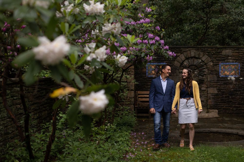 A couple walks through a garden with blooming white and purple flowers against a stone wall.