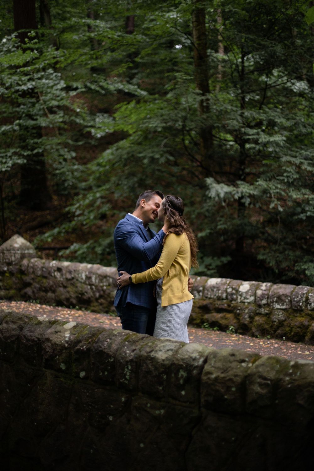 Romantic couple embracing on stone bridge surrounded by lush forest.