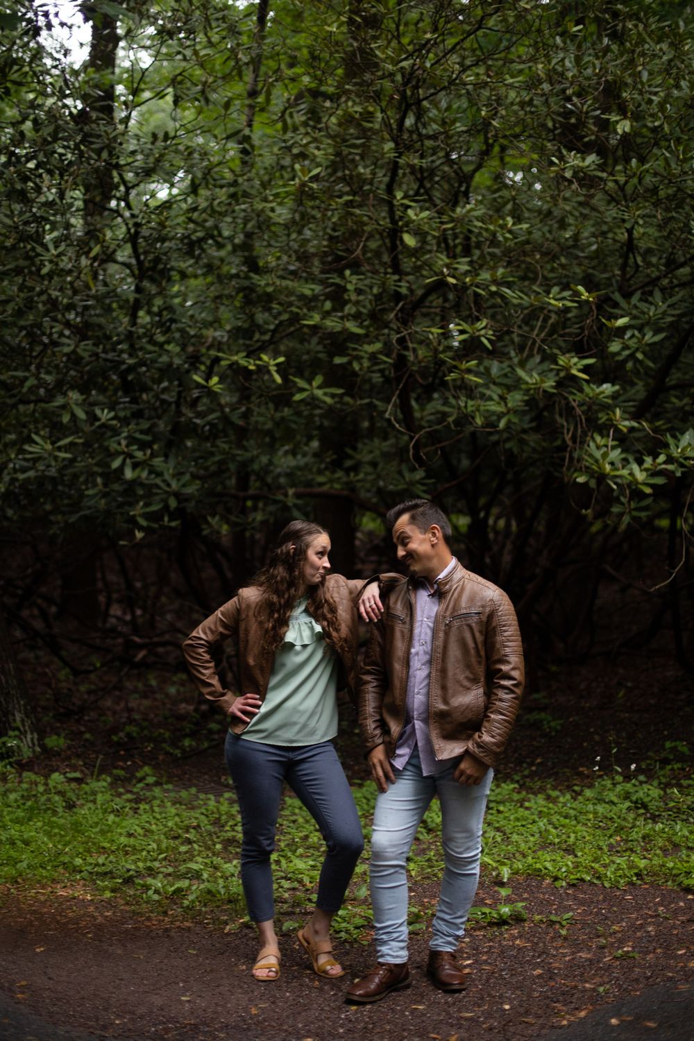 A romantic moment between a couple wearing matching brown leather jackets on a forest path.