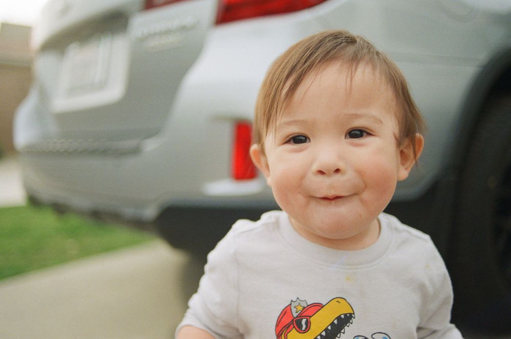 Toddler in white t-shirt smiling at camera in outdoor setting.